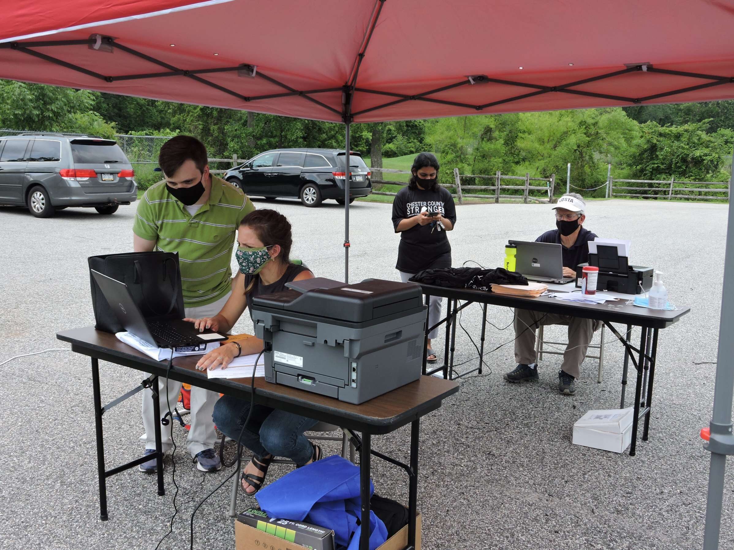 Four people are seen outside in a parking lot under a red tent pop-up tent. In the background, the trees are green. Two of the people, one man, one woman, sit at two different tables, each with a laptop and a printer visible. The other two stand next to the people at the table, one at each. They appear to be involved in conversations.