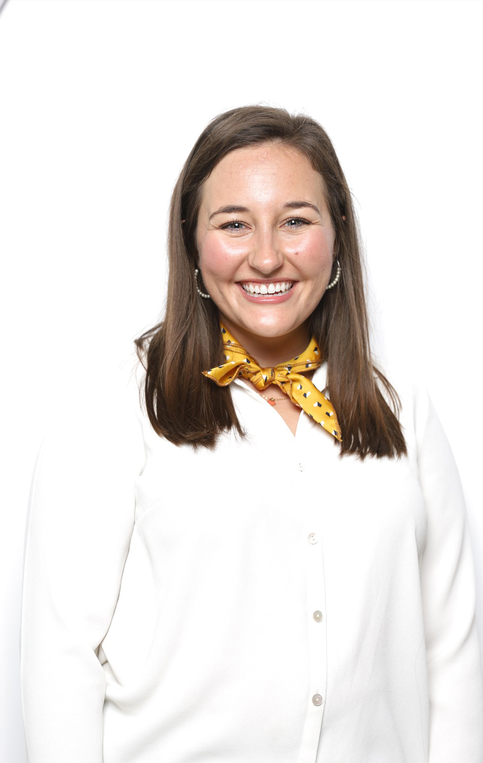 A white woman smiles for a portrait. She is wearing light-colored clothing and is standing against a white backdrop.