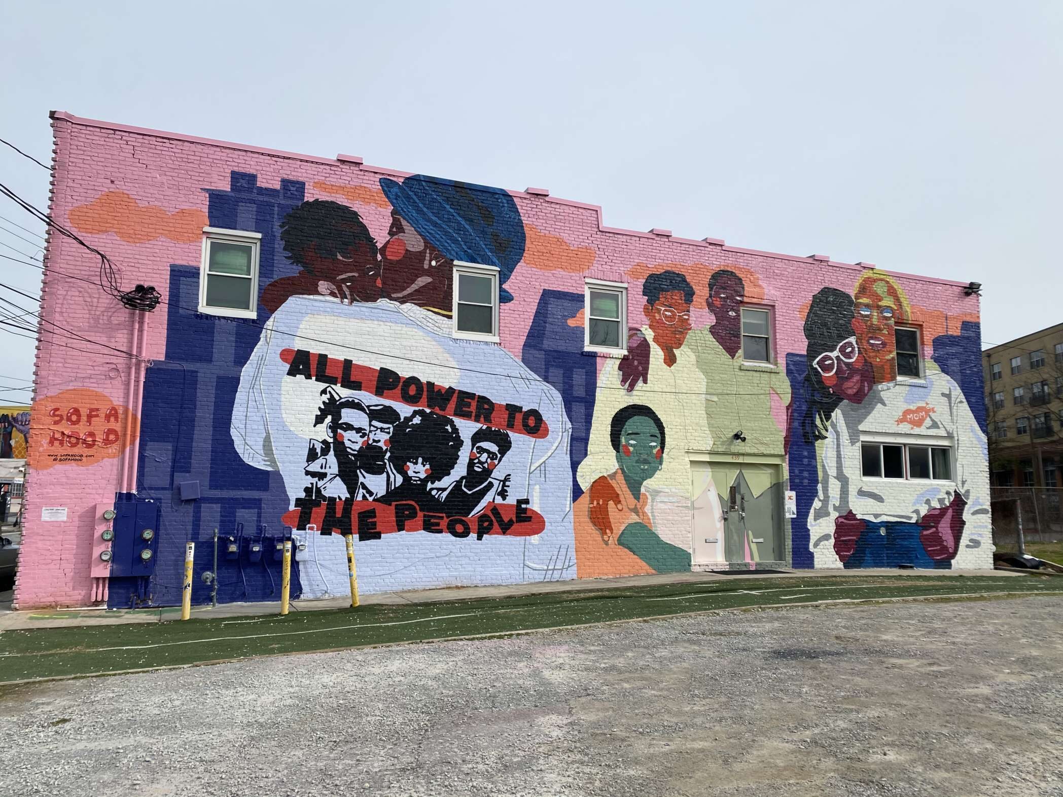 A colorful mural on the side of a building depicts abstracted black and brown people in living in an urban environment. One of the figures depicted on the mural wears a shirt showing Black Panther members. The shirt reads "All Power to the People."