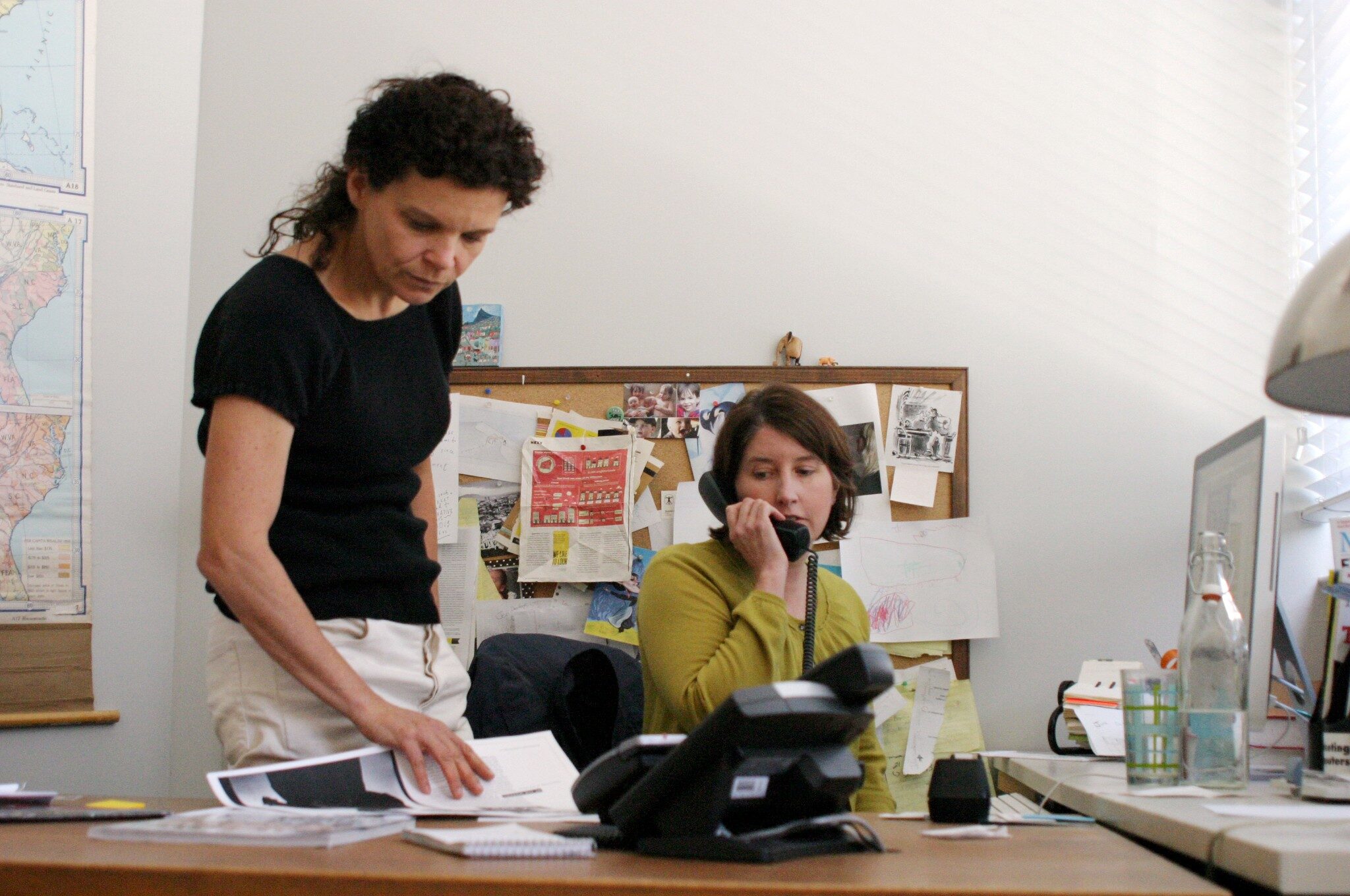 Two white women are seen in an office. One has a phone to her ear. The other examines papers on a desk.
