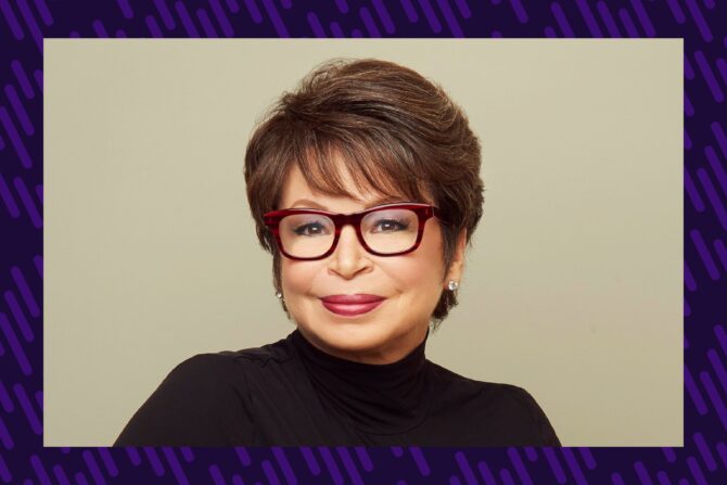 A woman in a black turtleneck smiles at the camera. She has short hair and dark purple glasses.