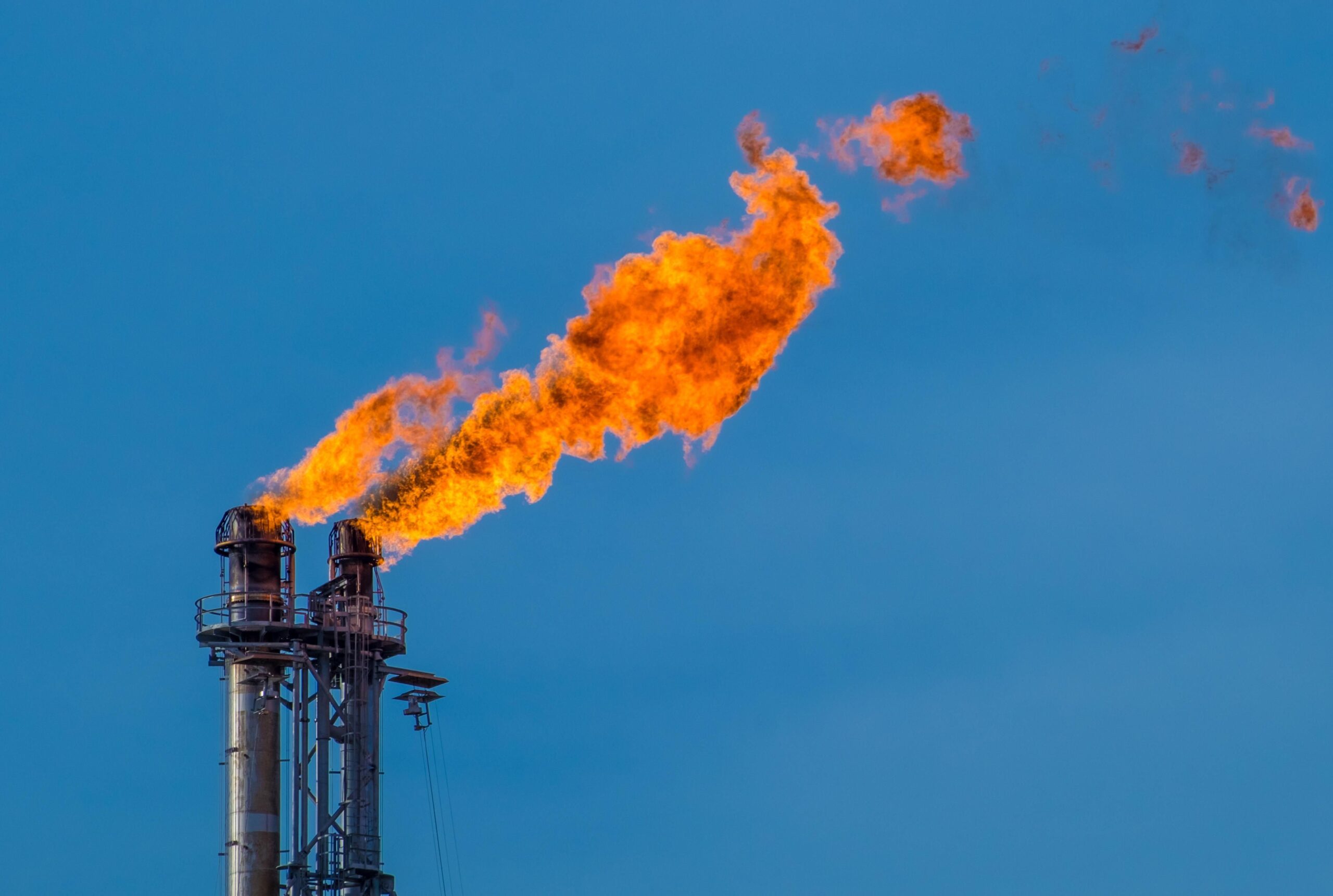 Large flames emit from the top of two large industrial metal chimneys, contrasting sharply with the bright blue-sky background.