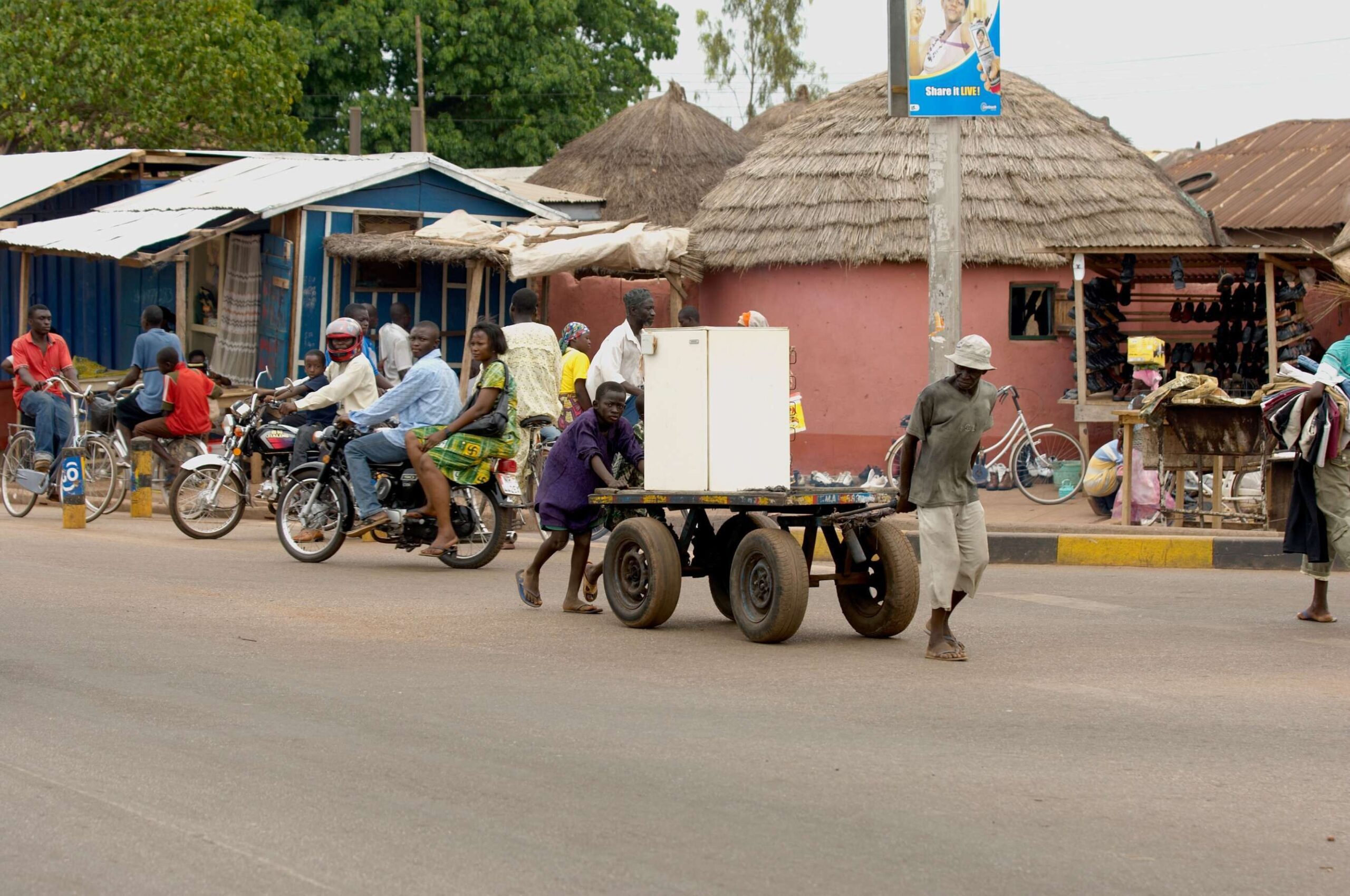 A man pulls refrigerator on cart on street in Tamale Ghana with a child helping. In the background, people walk through an urban landscape of one-story buildings with a mix of thatched roofs and metal roofs.