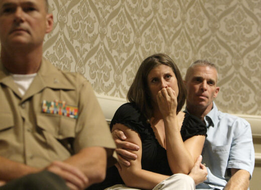 A woman with a pensive expression, hand to her mouth, is comforted by a man in a blue shirt, while a blurry military officer sits in the foreground.