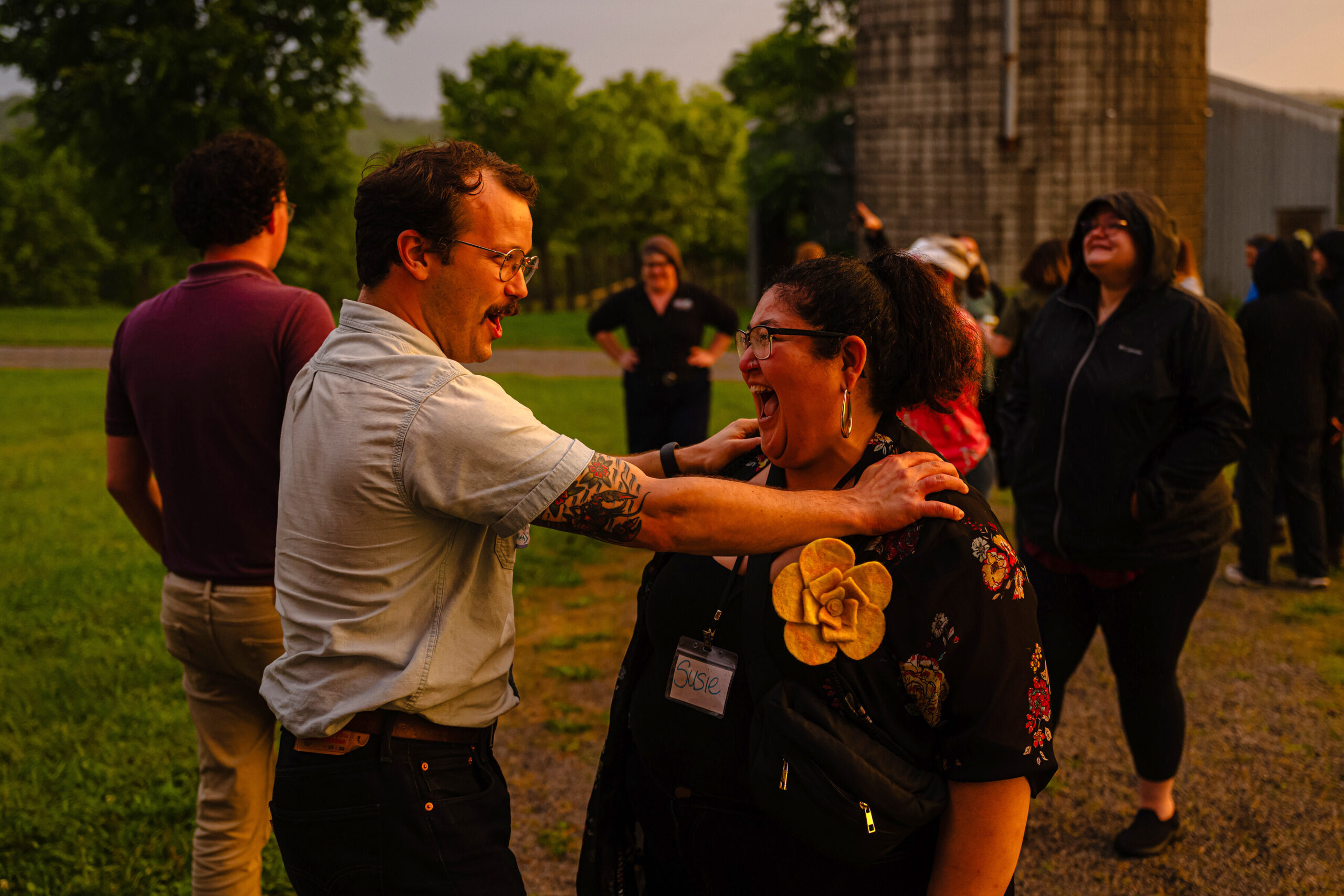 A man with a mustache and glasses, wearing a light shirt, laughs loudly while placing a hand on the shoulder of a woman with dark curly hair and glasses, who is also laughing with her mouth wide open. They are outdoors with other people and green trees in the background.