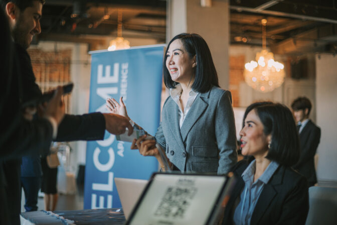 Three people at an event registration desk. A smiling woman in a gray suit receives badges, a man holds a phone, and another woman sits near a laptop displaying a QR code.
