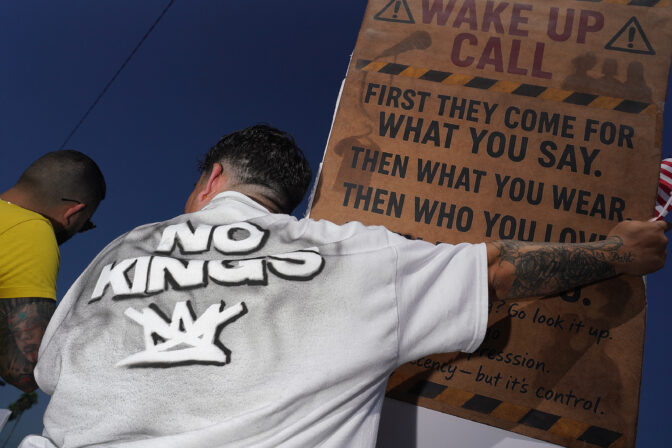Two men seen from behind; one wears a "No Kings" shirt and holds a brown sign that reads "WAKE UP CALL" and "FIRST THEY COME FOR WHAT YOU SAY. THEN WHAT YOU WEAR. THEN WHO YOU LOVE." He also holds a small American flag.