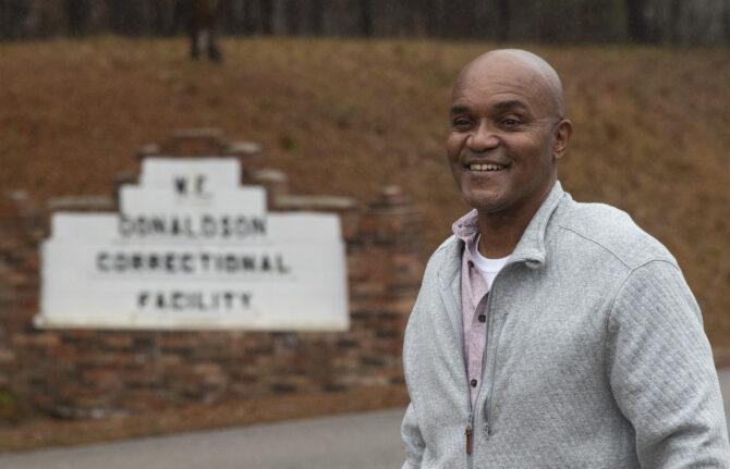 A smiling bald Black man in a grey jacket stands in front of a blurred sign for W.E. Donaldson Correctional Facility.