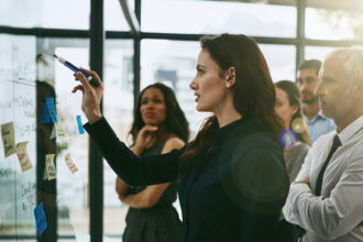 A woman writes on a glass whiteboard covered with notes and diagrams while a small group of colleagues stands behind her, watching and collaborating in a modern office setting.