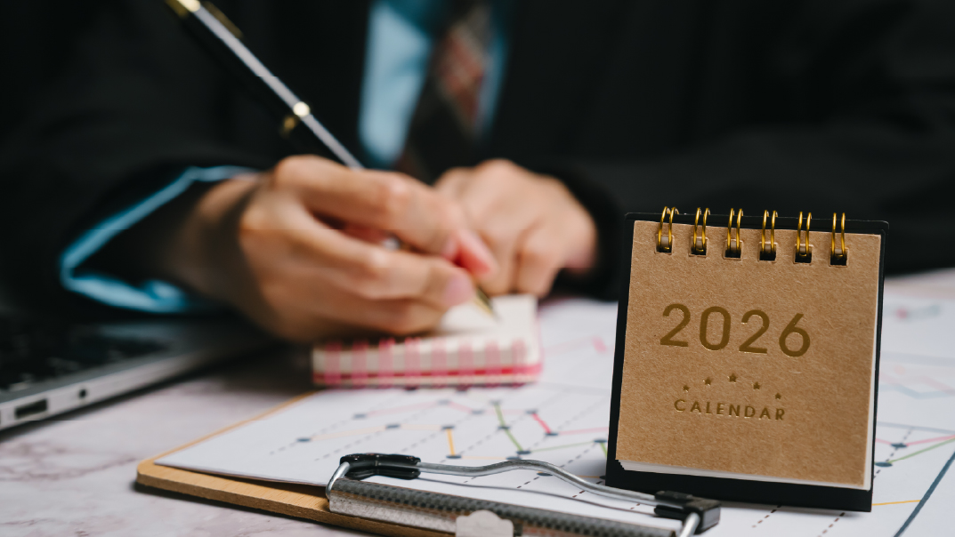Person in a business suit writing in a small notebook at a desk, with a 2026 desk calendar in the foreground and charts on a clipboard nearby.