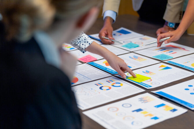 Paperwork and group of peoples hands on a board room table at a business presentation or seminar. The documents have financial or marketing figures, graphs and charts on them. People are pointing at the data