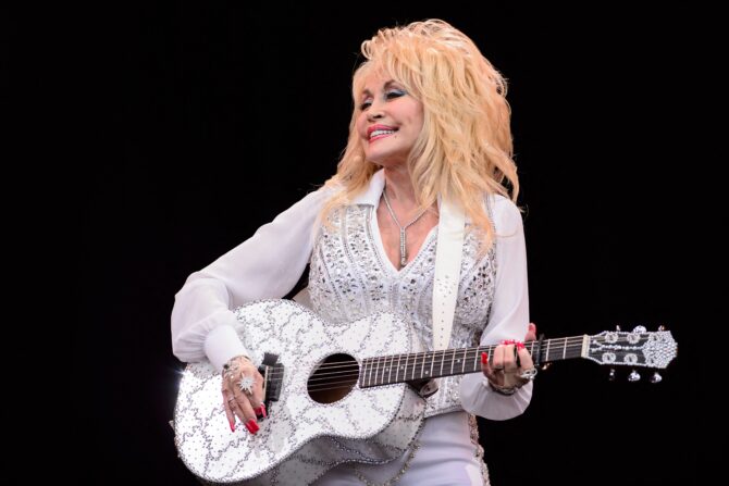 Dolly Parton performs onstage with a guitar; dressed in all white.
